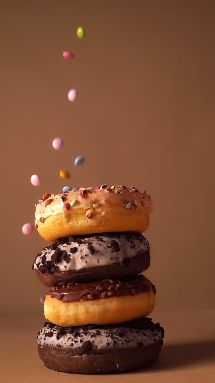 Em câmera lenta, donuts empilhados com cobertura de baunilha, glacê de chocolate e migalhas de biscoito, enquanto pedaços de chocolate coloridos confeitados caem de cima. Fundo de estúdio bege quente.
