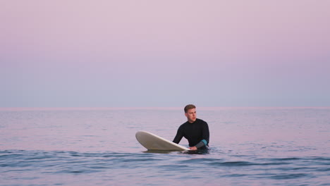 man wearing wetsuit sitting and floating on surfboard at sea as waves break around him