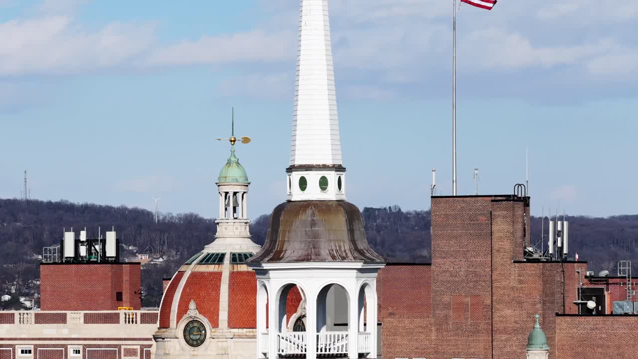 Vista aerea di York, Pennsylvania in inverno con tetti storici, campanili di chiese e una bandiera americana che sventola sotto un cielo azzurro e una luce solare soffusa. Inquadratura ampia. Alberi della foresta senza foglie in lontananza.