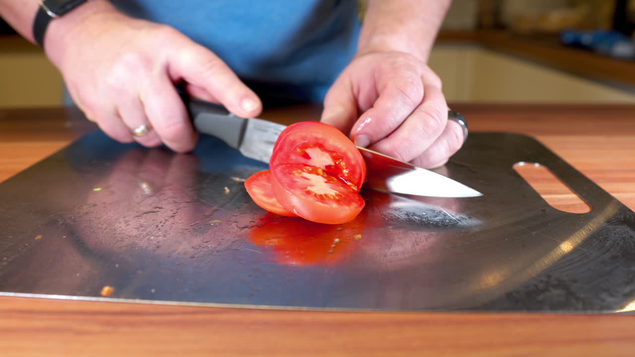Close-up of a person slicing a fresh tomato on a stainless-steel cutting board in a kitchen. The hands carefully hold a sharp kitchen knife while cutting tomato slices on a wooden countertop
