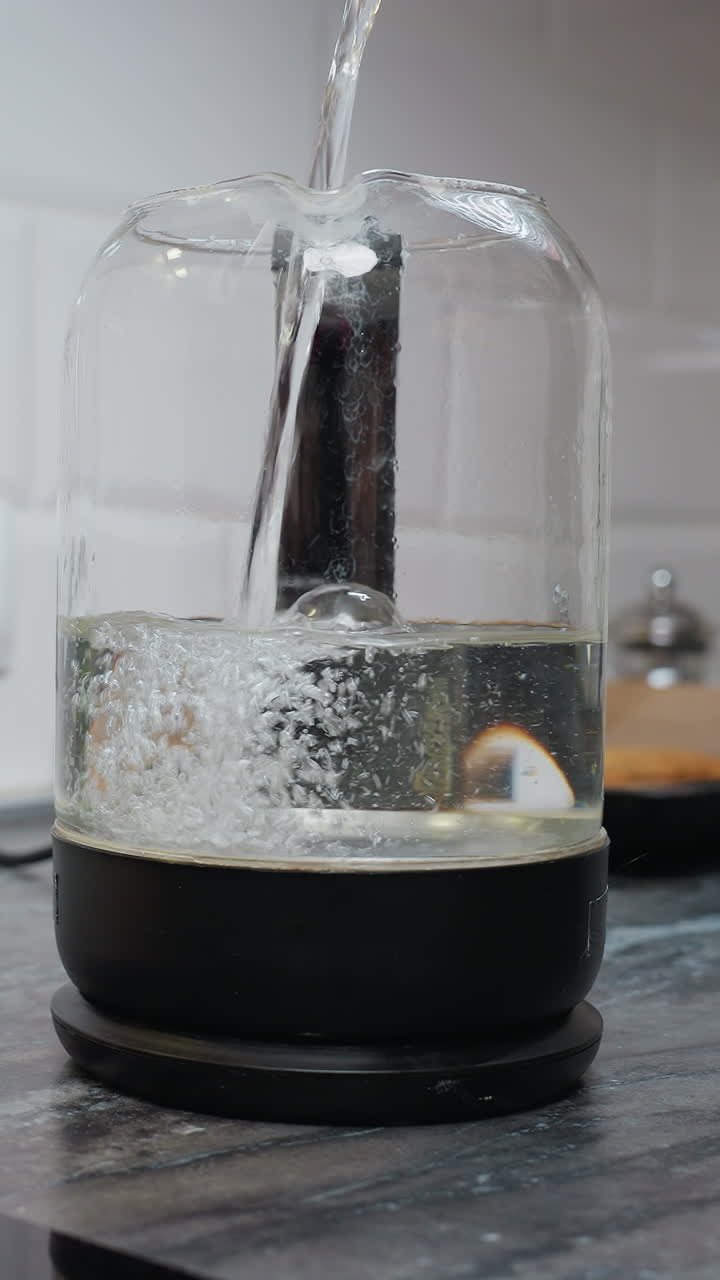 부 카운터에 있는 유리 항아리에 물을 부어 넣고 있다. 흐릿한 배경에는 벽에 있는 소켓, 구운 물품이 있는 트레이, 컵, 장식용 구슬이 있다.