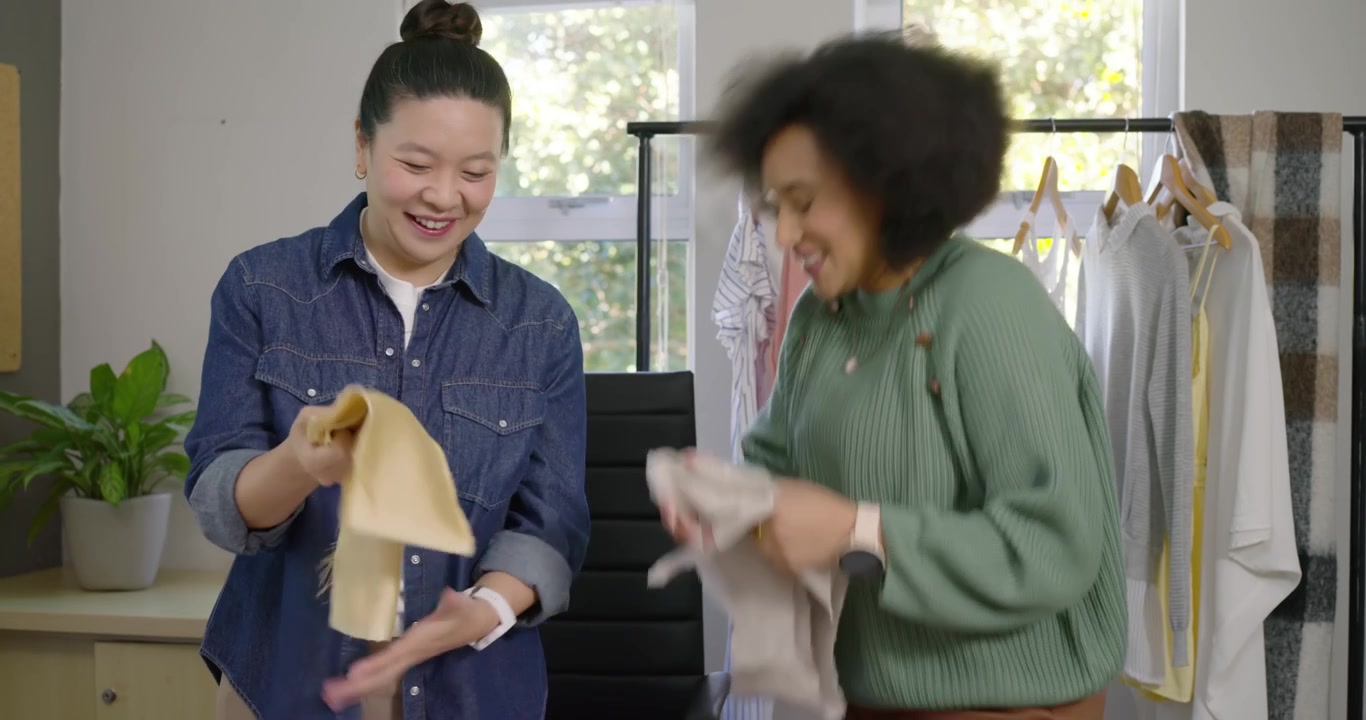 Diverse female stylists comparing fabrics, denim woman extending knit sample with tablet, choosing