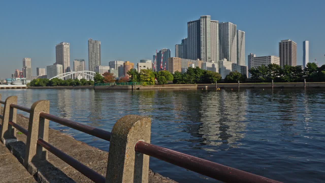 A panoramic view of Tokyo's modern skyline from the waterfront of Hamarikyu Gardens, featuring high-rise buildings and reflections on the calm water.