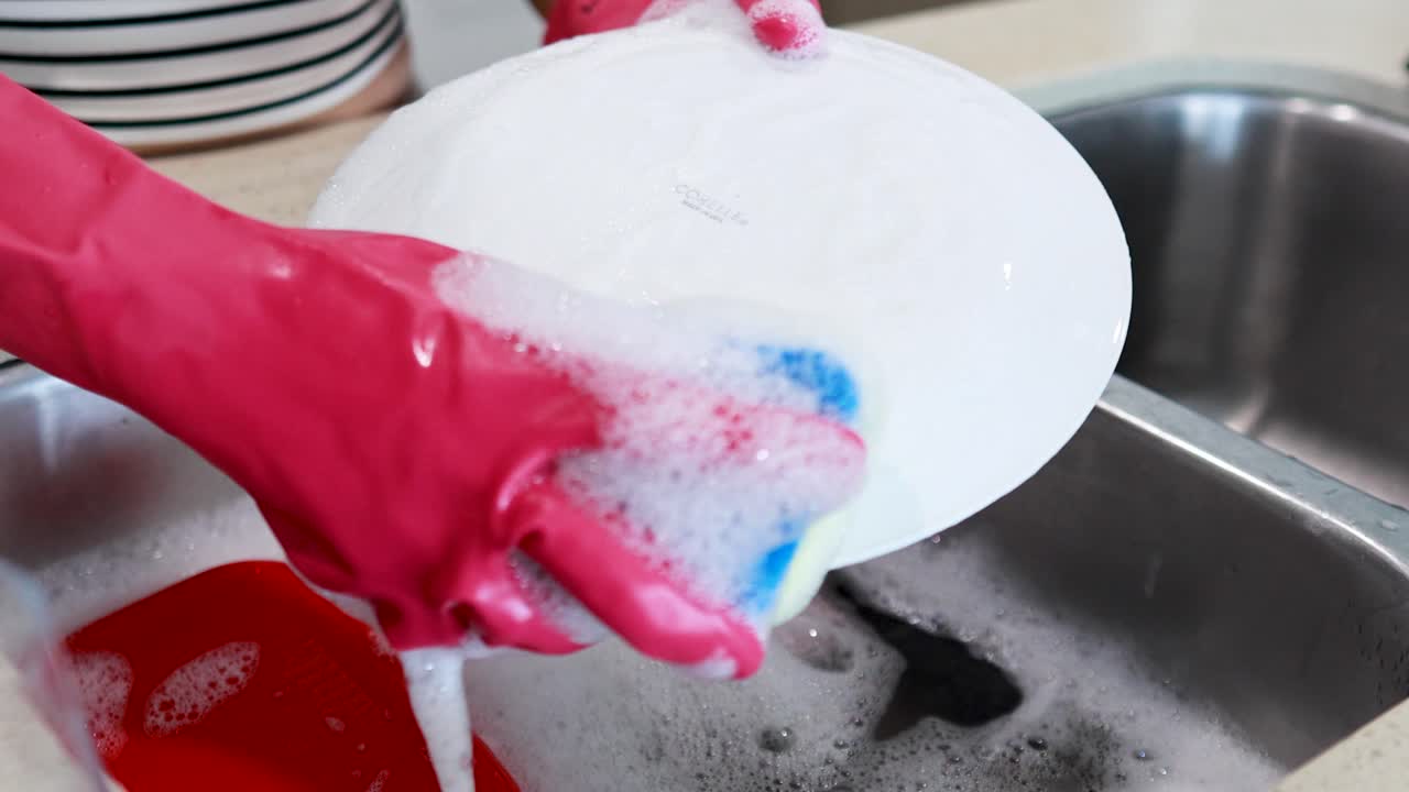 Hands in pink gloves washing a white plate with a sponge in a kitchen sink filled with soapy water