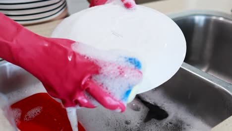 hands in pink gloves washing a white plate with a sponge in a kitchen sink filled with soapy water