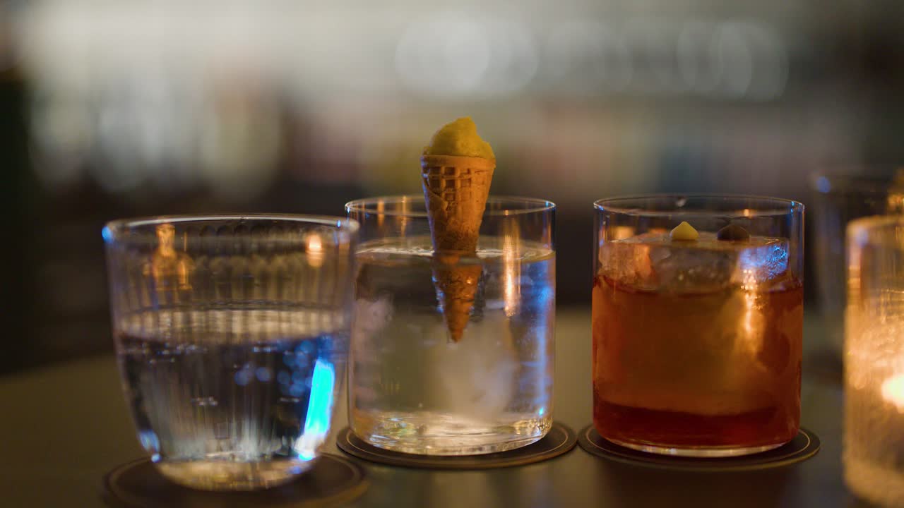 Three cocktails with ice and garnish on bar table, evening bokeh lighting, static camera shot