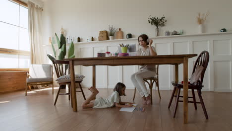 wide shot of a mother making a videocall while her daughter is distracted under the table drawing