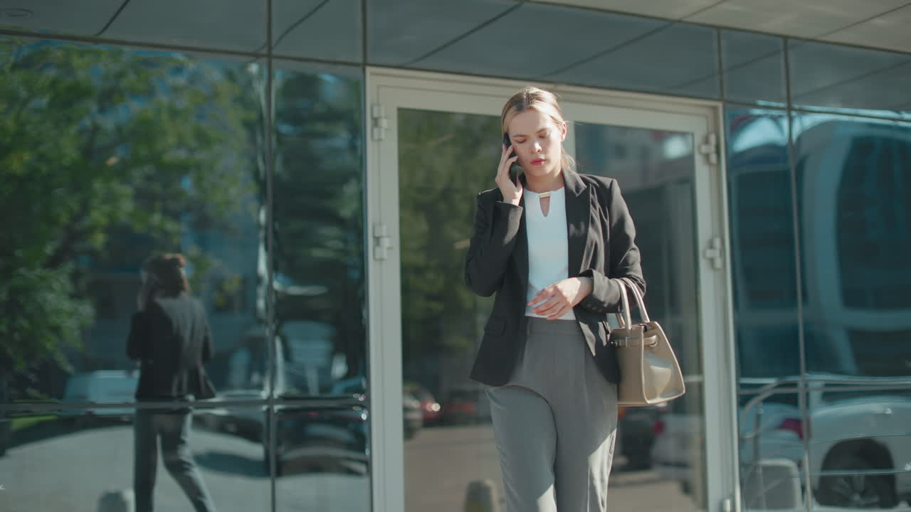 Lawyer exits mall on phone call with attentive expression, dressed in professional attire, holding handbag while walking, urban glass building reflects cityscape and vehicles