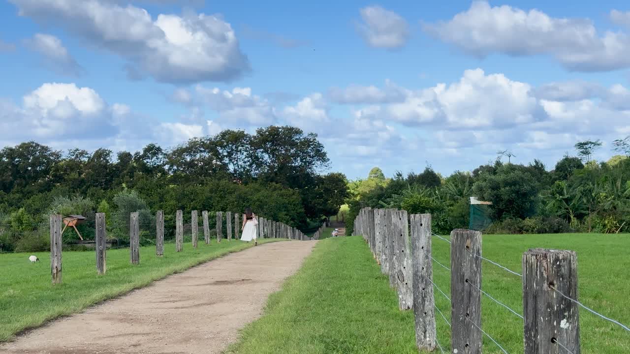 A woman walks down a sunlit dirt path bordered by wooden posts and green grass in a rural garden, with steady camera movement and natural daylight
