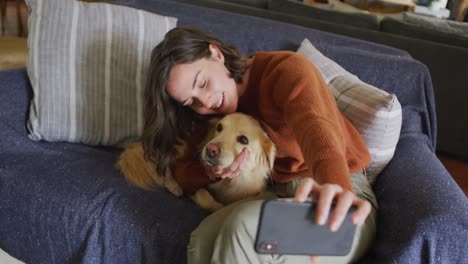 smiling caucasian woman taking selfie with smartphone and stroking her pet dog at home