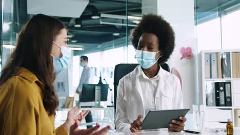 vista de cerca de una doctora afroamericana con mascarilla médica sosteniendo una tableta y explicando a una paciente el tratamiento del coronavirus en consulta médica