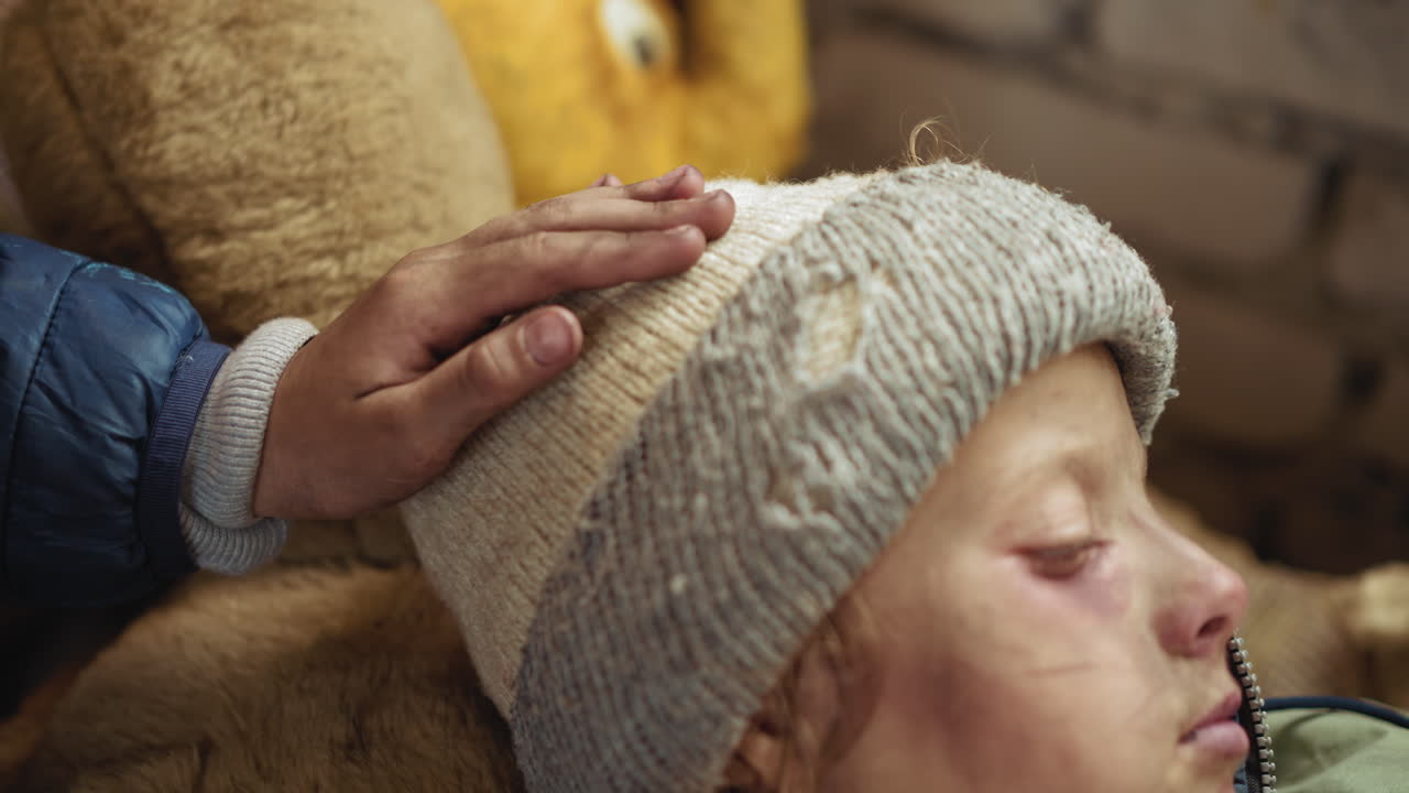 차가운 방 안 침구에 누워있는 낡은 니트 모자를 쓴 멍든 아이의 머리를 부드럽게 두드리기 위해 뻗은 아이 손의 클로즈업; 다정한 손길은 병, 위로, 보호와 조용한 보살핌을 암시해요