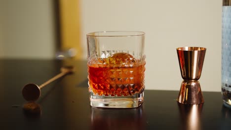 Negroni-Cocktail-with-spherical-ice-front-view-on-top-of-a-bar-with-jigger-and-mixing-stick