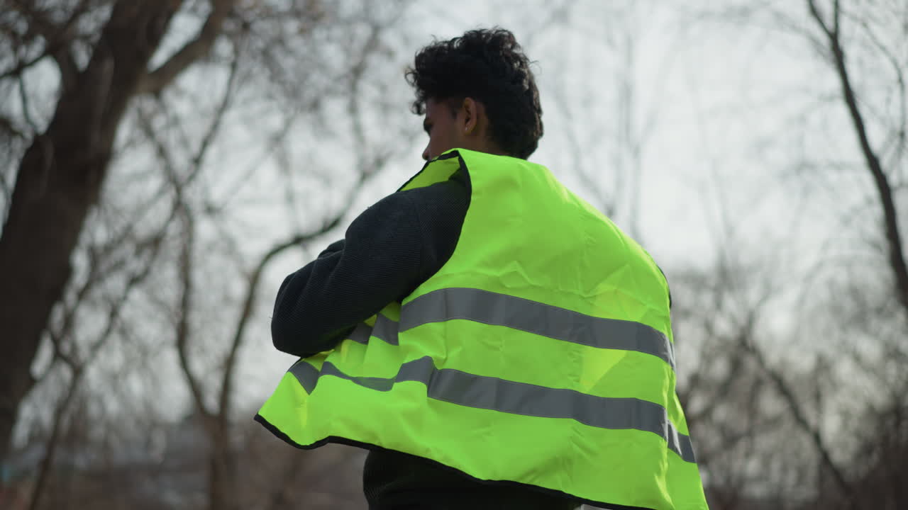 Man outdoors putting on bright neon green reflective safety vest, lifting arm over head to adjust fit, preparing for work or volunteer activity in open park area with dry grass during sunny day