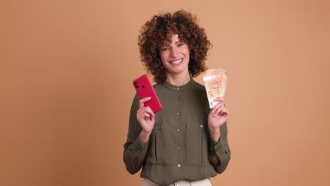 excited woman with cash and mobile phone in beige studio