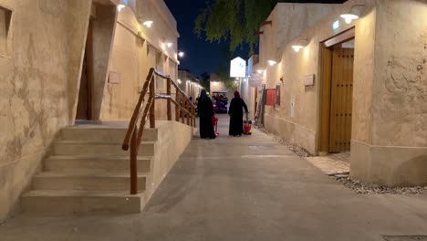 Una-Familia,-Mujeres,-Niños,-Niños,-Caminan-En-El-Casco-Antiguo,-En-El-Centro-De-Doha,-Qatar,-Un-Camino-A-Pie-Con-Una-Casa-De-Construcción-De-Adobe-De-Adobe,-Una-Puerta-De-Madera-De-Diseño-Y-Una-Luz-Cálida-Que-Ofrecen-Cultura-Tradicional