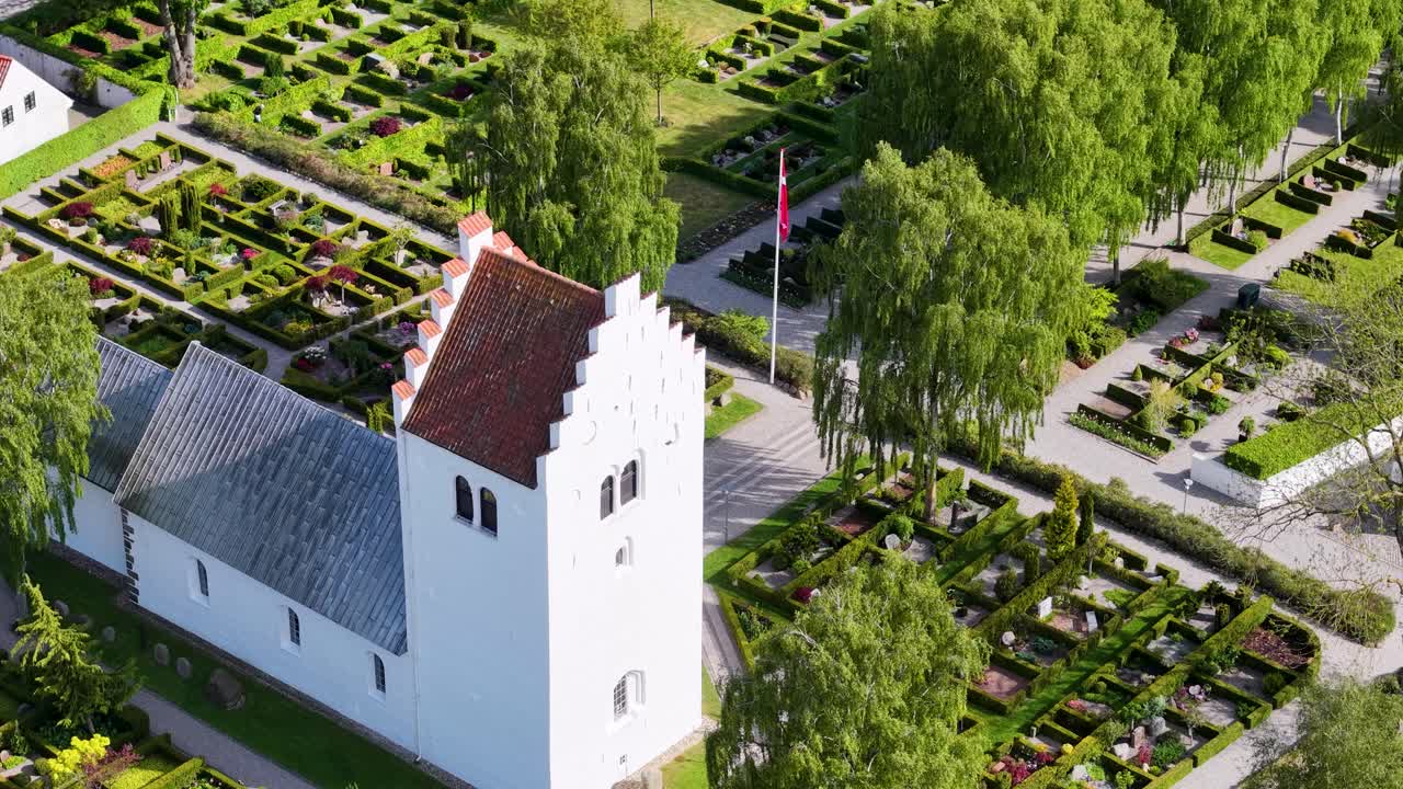 Aerial drone footage of a traditional white church with a red roof surrounded by a green cemetery and trees in the countryside