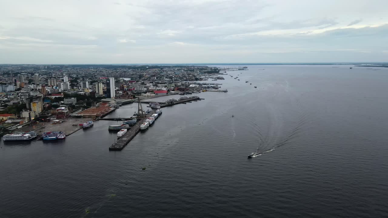 Fiume nero a Manaus mentre si sorvola una barca, con la città sulla sinistra e un orizzonte lontano in una giornata nuvolosa. Ripresa aerea ampia e in avanti dall'alto registrata in 4K Apple ProRes 422