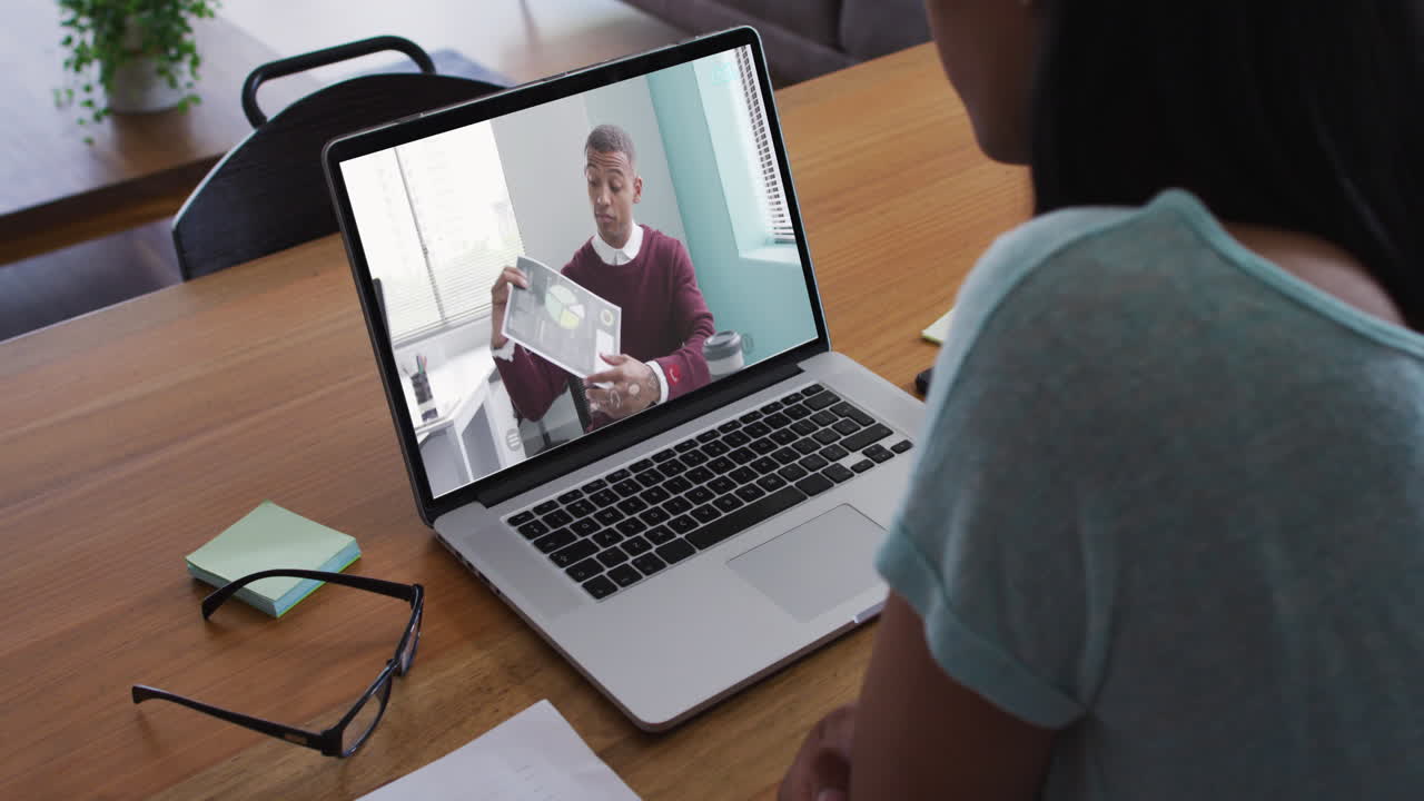 mujer de negocios biracial sentada en el escritorio usando una computadora portátil haciendo una llamada de video con un colega masculino