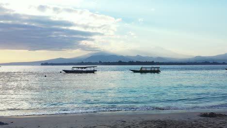 long boats floating in the peaceful sea, sandy beach and hills in the background covered in the clouds at sunset