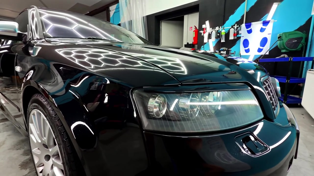 Auto nera lucida in uno studio di detailing interno, con riflessi di luce intensa sul cofano, sul faro e sulla carrozzeria anteriore lucida.