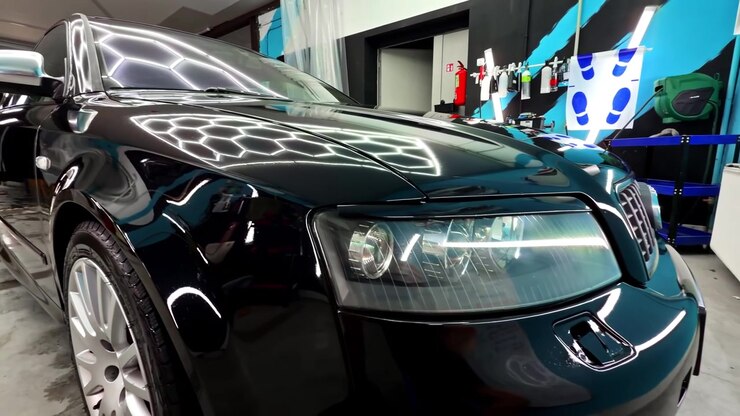 Auto nera lucida in uno studio di detailing interno, con riflessi di luce intensa sul cofano, sul faro e sulla carrozzeria anteriore lucida.