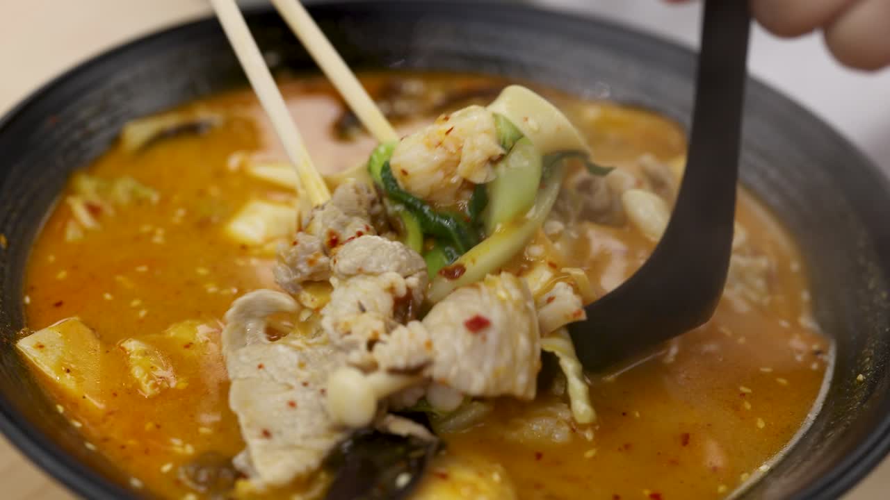 Chopsticks and ladle lift pork, vegetables, mushrooms from steaming red hotpot soup in close-up