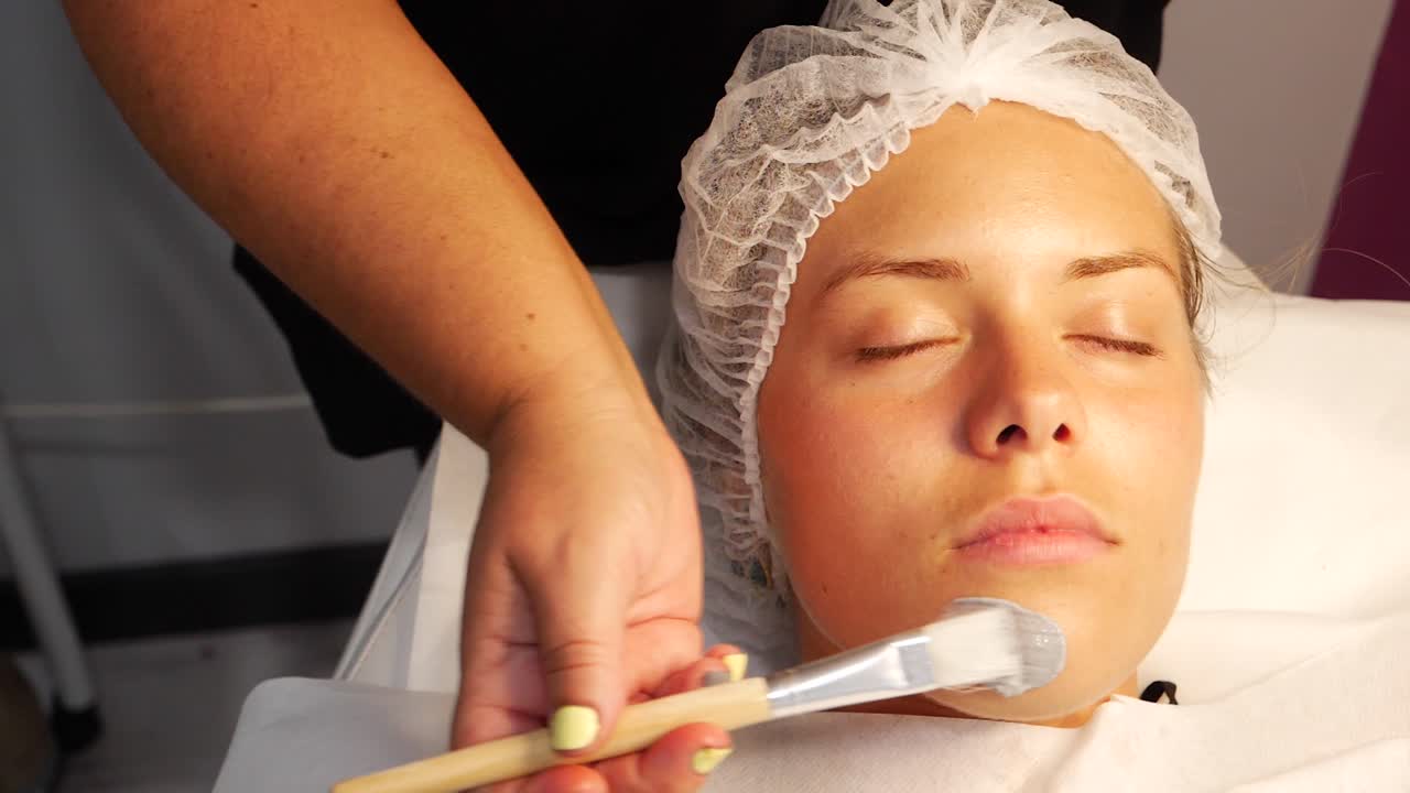 Stable handheld frame-right close-up headshot of a woman having a facial done by a professional esthetician whose right hand holds a brush with the skincare product