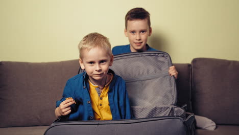 cheerful brothers playing at home with grey valise on the sofa. a boy opens the big suitcase and a little boy gets out inside of it and eat something.