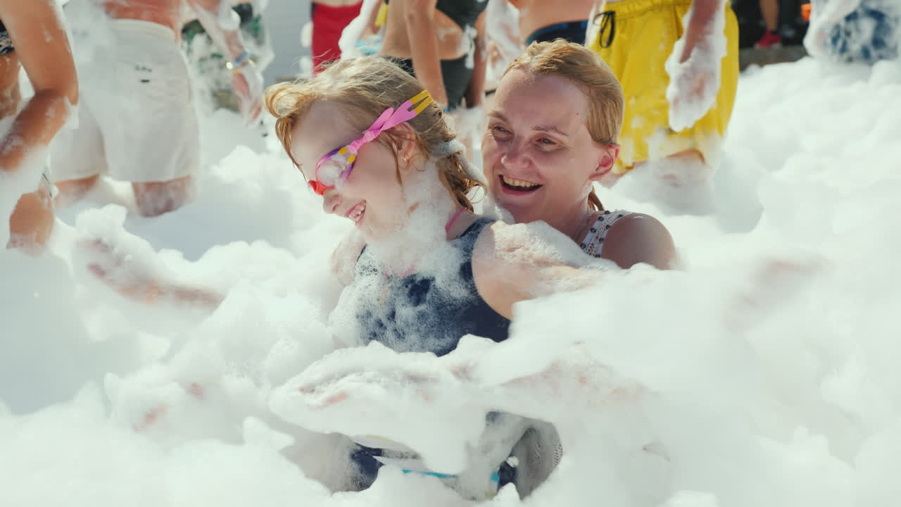 mamá y su pequeña hija juegan con jabón y espuma en una multitud de turistas en una fiesta en la playa en un res