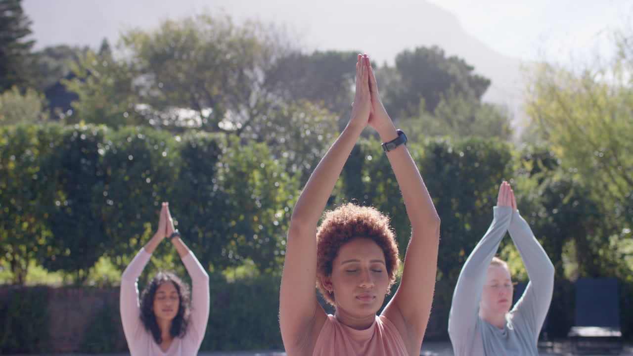 Practicing yoga outdoors, diverse female friends meditating with hands raised in park