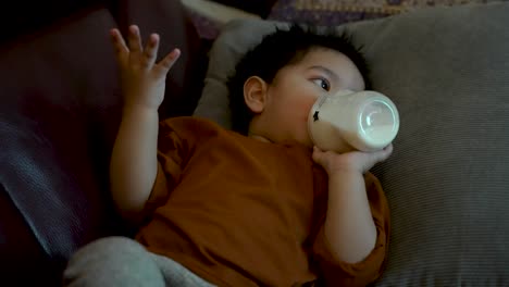 Toddler-in-orange-shirt-lying-down-drinking-from-bottle,-focused-on-TV-screen,-indoors