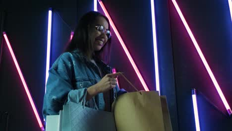 young girl in blue denim jacket with shopping bags on neon lamps background.