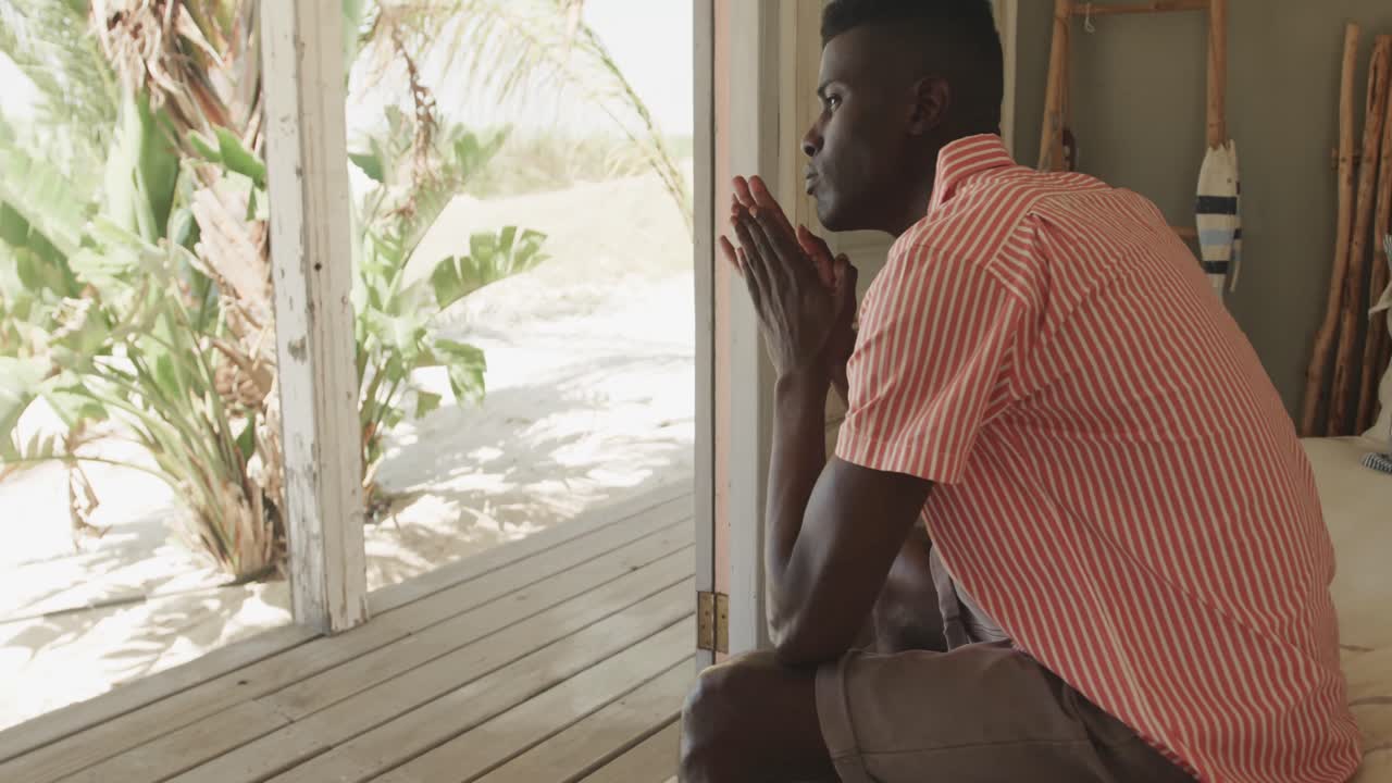 Uomo afroamericano pensieroso seduto sulla soglia di una casa al mare che guarda altrove, al rallentatore.