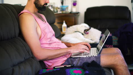 Bearded-Caucasian-man-at-home-on-sofa-typing-on-notebook-with-phone-to-the-side,-medium-shot