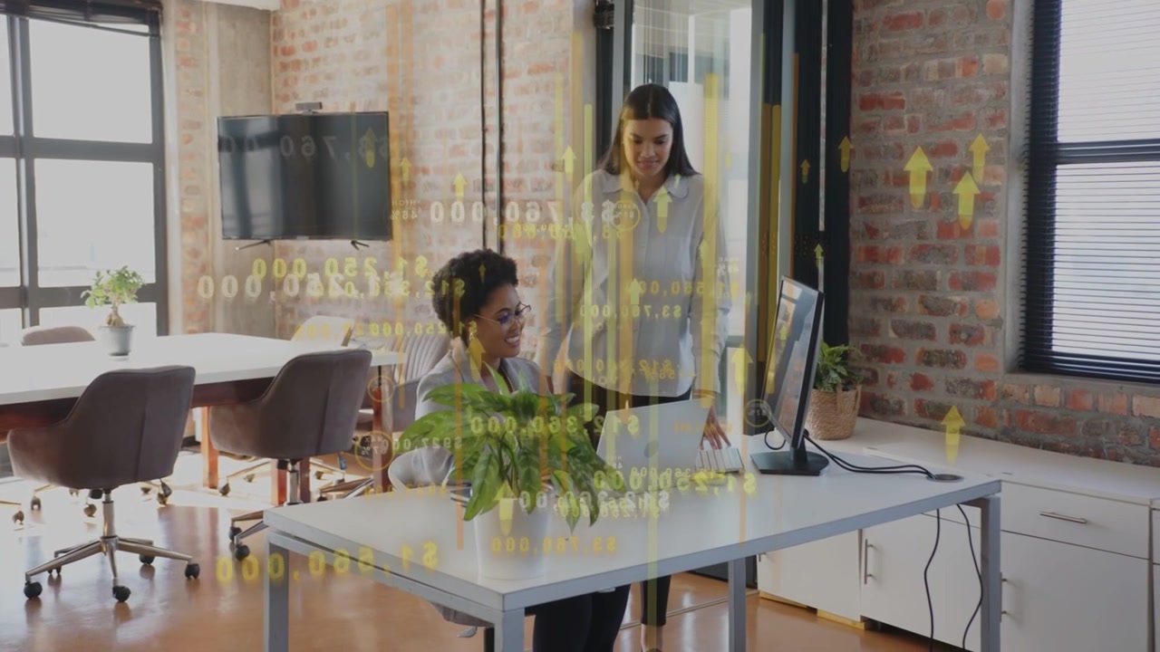 Seated woman typing laptop while colleague entering leaning and reviewing yellow business chart