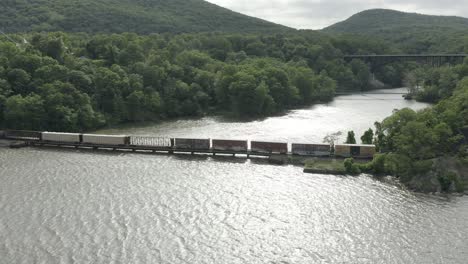 Drone-shot-of-a-long-cargo-train,-flying-to-the-left-and-panning-right,-with-the-train-going-over-a-river