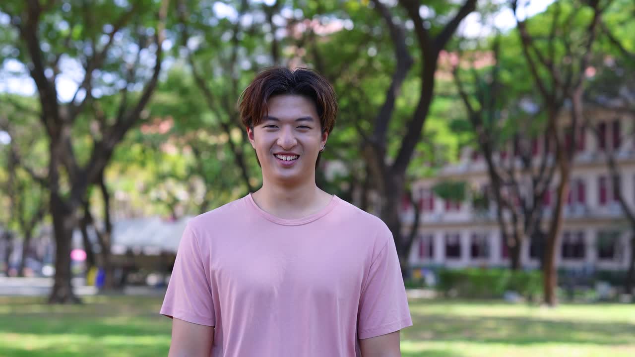 ピンクのシャツを着た若いアジア人男性が、キャンパスの敷地で屋外で笑顔で笑っています。