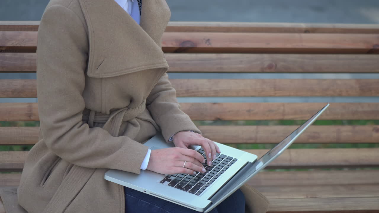 Woman in a brown coat working on her laptop on a bench in the park