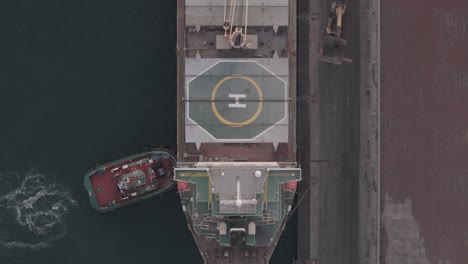 aerial shot following a cargo ship from bow to stern, featuring a helipad, grain containers, and tugboats assisting as it docks at the port.
