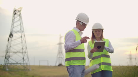 Ingeniería-Trabajando-En-Una-Torre-De-Alto-Voltaje-Verifique-La-Información-En-La-Tableta-Dos-Empleados,-Hombre-Y-Mujer