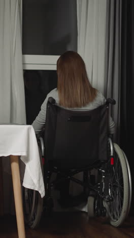 pensive woman sits in wheelchair near kitchen table looking out window. young housewife with cerebral palsy spends time alone overthinking backside view