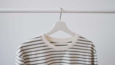 woman's hand picks up white wooden hanger with striped vest, jacket, sweatshirt against white wall. minimalistic clothing concept, store advertising, sale, discounts, shopping