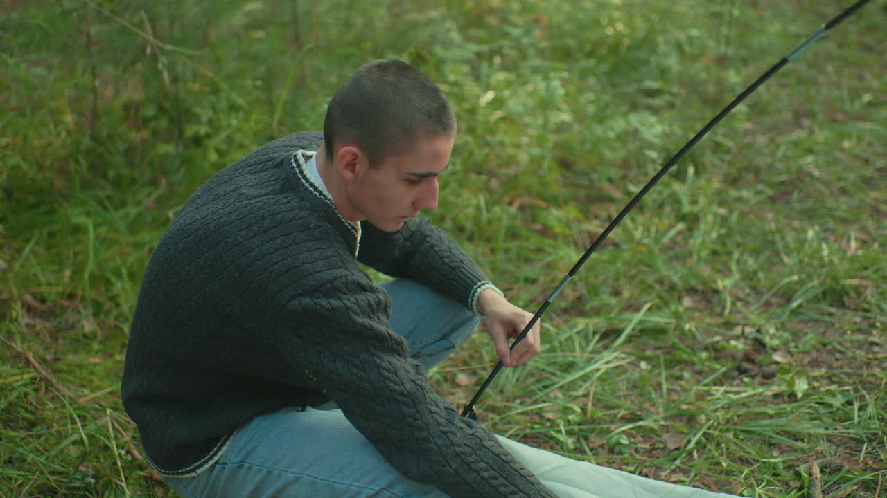 Focused researcher in sweater squats on forest grass adjusting tent pole into ring of fabric while setting up camping site surrounded by greenery and soft daylight in tranquil outdoor setting