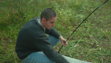 focused researcher in sweater squats on forest grass adjusting tent pole into ring of fabric while setting up camping site surrounded by greenery and soft daylight in tranquil outdoor setting