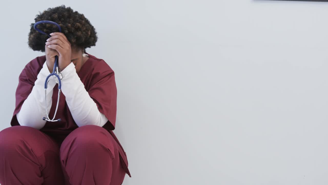 Tired african american female doctor squatting in hospital corridor with copy space, slow motion