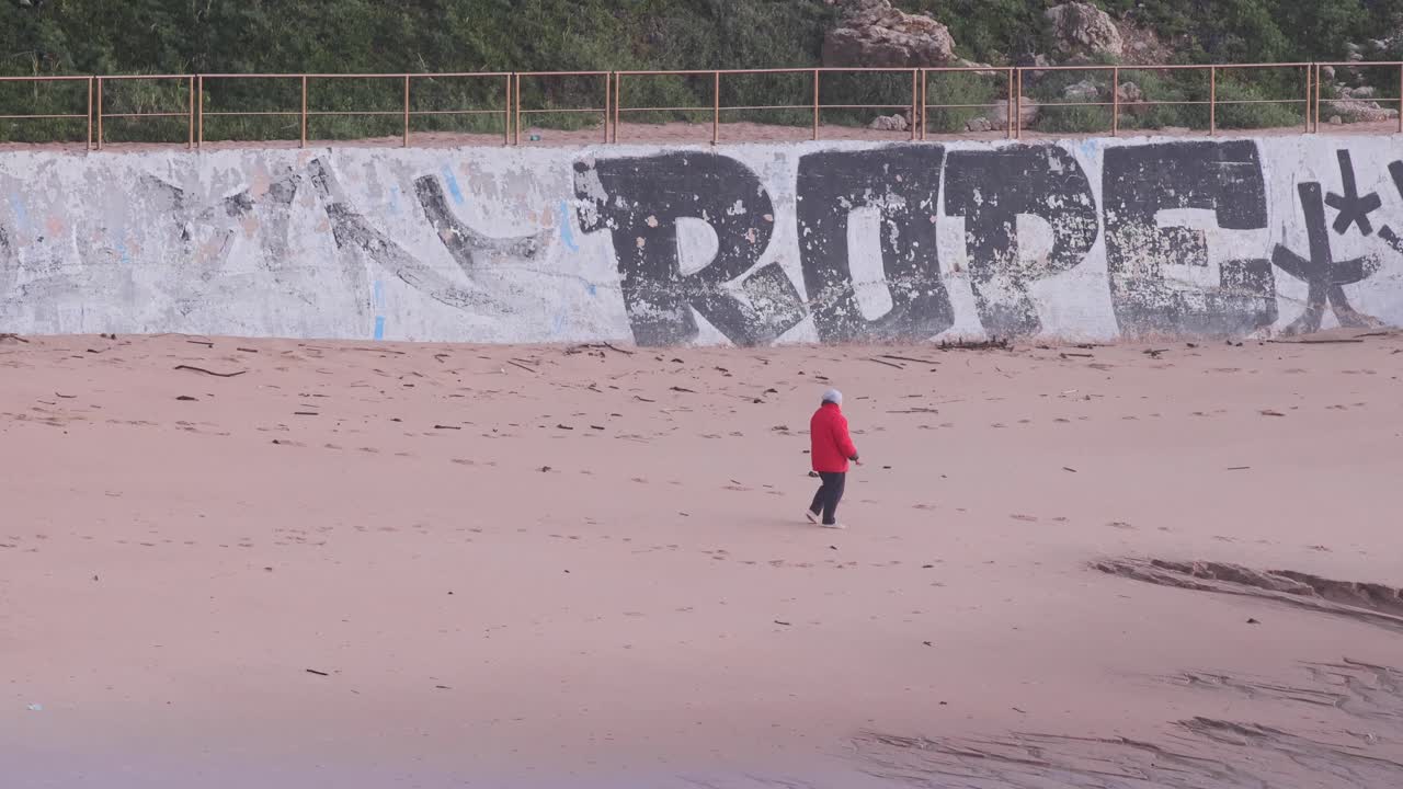 vista trasera de una mujer mayor con chaqueta roja caminando por la playa