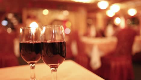 two glasses of red wine on a table in a softly lit restaurant as a waitress walks past, with a shallow depth of field and warm ambiance