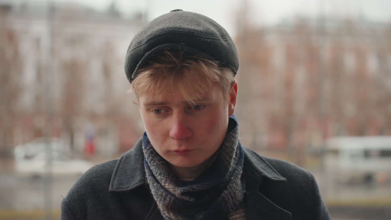 Young white creator in cap thinking, rainy urban sidewalk, scarf and coat, downward glance, planning project, checking notes mentally, film student energy, cinematic mood, subtle background bokeh