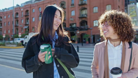 young cheerful women walking on city street with coffee and talking